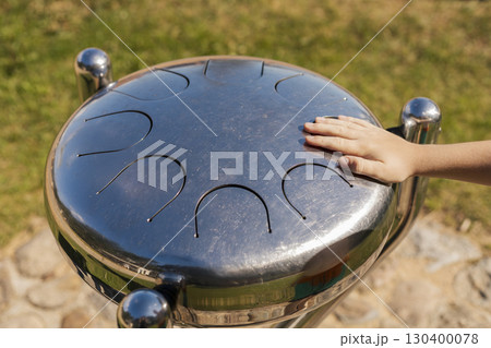Hand rests on the metallic surface of a musical instrument while a child enjoys creating sounds in a vibrant park under clear blue skies in the afternoon Hand rests on the metallic surface of a musical instrument while a child enjoys creating sounds in a vibrant park under clear blue skies in the afternoon 130400078