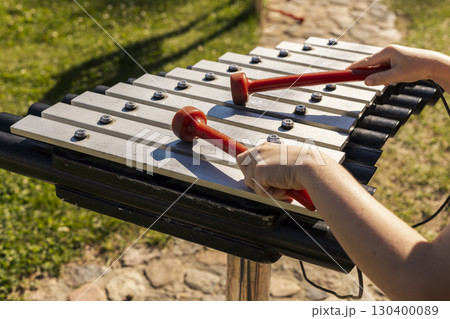 Children are having fun creating melodies on a xylophone with bright mallets in a lively outdoor park. The sun illuminates their joyful expressions and the surrounding green grass 130400089
