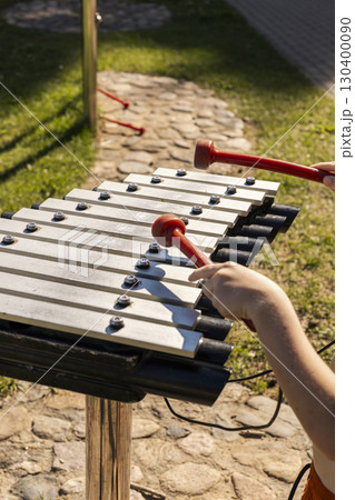 Children are having fun creating melodies on a xylophone with bright mallets in a lively outdoor park. The sun illuminates their joyful expressions and the surrounding green grass Children are having fun creating melodies on a xylophone with bright mallets in a lively outdoor park. The sun illuminates their joyful expressions and the surrounding green grass 130400090
