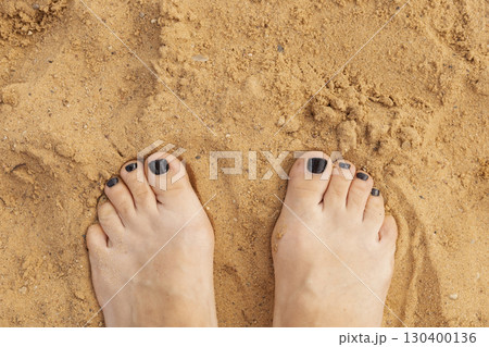 Toes with black nail polish resting on golden sand, capturing the essence of a sunny summer day at the beach. Feelings of relaxation and enjoyment abound in this shoreline setting 130400136