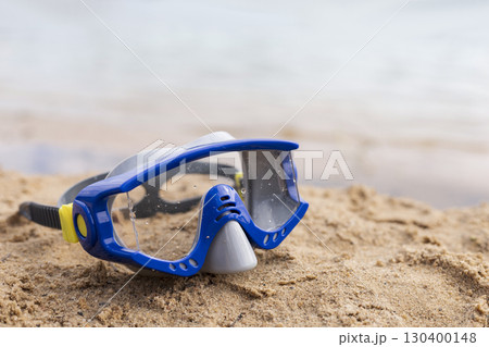 A blue diving mask sits on warm sand at a beach during sunset. Gentle waves lap the shore, creating a peaceful ambiance perfect for water activities A blue diving mask sits on warm sand at a beach during sunset. Gentle waves lap the shore, creating a peaceful ambiance perfect for water activities 130400148