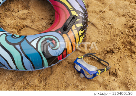 A vibrant swimming tube lies on golden sand beside a pair of snorkel goggles. The bright colors of the gear contrast with the warm beach setting under clear blue skies 130400150
