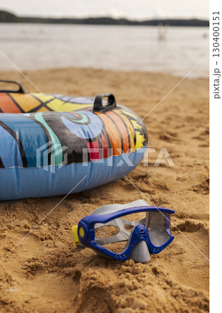 Bright inflatable float with a vibrant design rests on warm sand, next to a snorkel. Calm water and distant beachgoers create a relaxing summer atmosphere 130400151