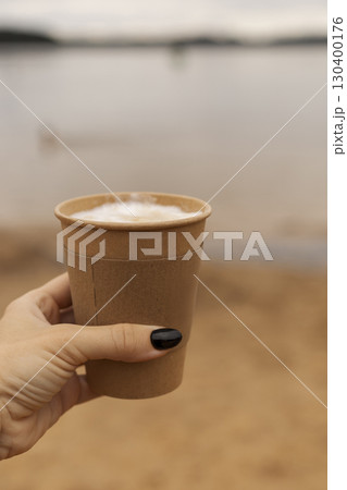 A hand with dark nails holds a brown paper cup filled with coffee near the water's edge of a tranquil lake in the late afternoon, with soft lighting and gentle waves A hand with dark nails holds a brown paper cup filled with coffee near the water's edge of a tranquil lake in the late afternoon, with soft lighting and gentle waves 130400176