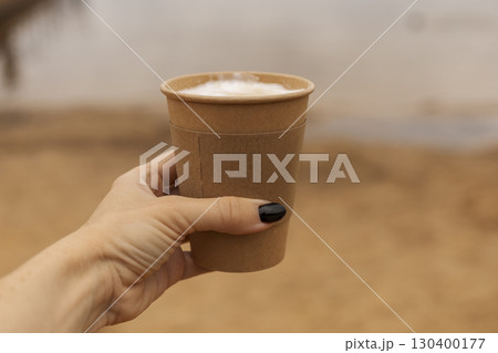 A hand holds a brown paper cup filled with a warm drink by a tranquil lakeside. The sandy shore and clear water create a relaxing outdoor atmosphere 130400177