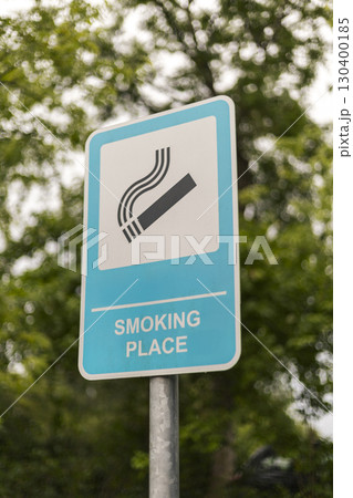 The sign shows a clear symbol and text indicating a smoking place, positioned among lush trees on a cloudy day, guiding smokers to the designated area The sign shows a clear symbol and text indicating a smoking place, positioned among lush trees on a cloudy day, guiding smokers to the designated area 130400185