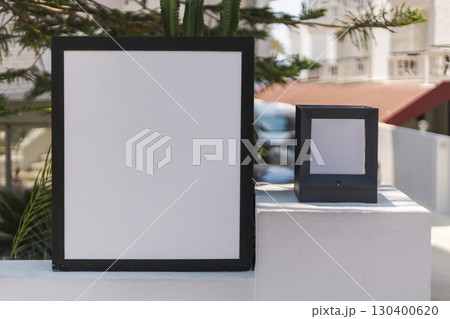 A blank sign rests on ledge surrounded by lush greenery. Bright sunlight illuminates the area, revealing a glimpse of an urban landscape in the background. Signboard mockup. 130400620