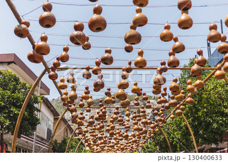 A lively street market showcases an array of hanging gourds suspended overhead, creating a festive atmosphere. Bright sunlight highlights the earthy colors of the gourds against a clear blue sky A lively street market showcases an array of hanging gourds suspended overhead, creating a festive atmosphere. Bright sunlight highlights the earthy colors of the gourds against a clear blue sky 130400633