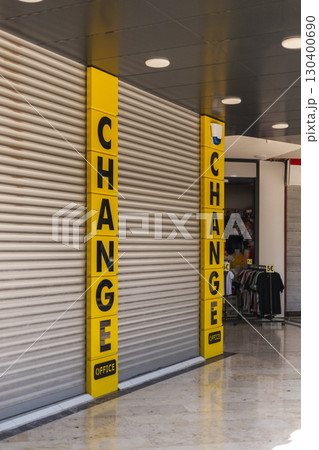 Bright yellow signs reading CHANGE indicate a currency exchange office. The shop is closed, with a metallic shutter down and other shops visible in the background 130400690