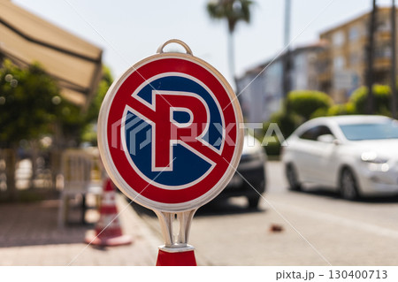 Bright day featuring a no parking sign prominently displayed near a street lined with vibrant greenery and parked cars. Palm trees create a tropical atmosphere in the background Bright day featuring a no parking sign prominently displayed near a street lined with vibrant greenery and parked cars. Palm trees create a tropical atmosphere in the background 130400713