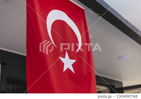 A vibrant red Turkish flag with a white star and crescent is hanging outside a modern building. The setting is lively, with bright sunlight shining down on the flag 130400748