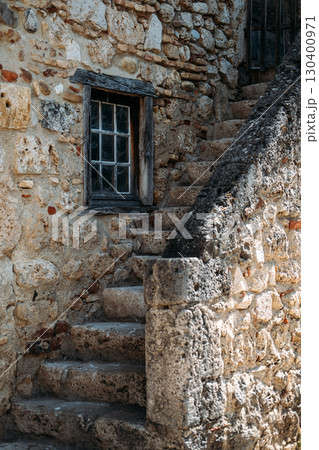 Aged stone staircase beside a rustic window framed by worn wooden beams on an old stone wall. Quiet living, countryside serenity, escapist domesticity, contemplative design Aged stone staircase beside a rustic window framed by worn wooden beams on an old stone wall. Quiet living, countryside serenity, escapist domesticity, contemplative design 130400971