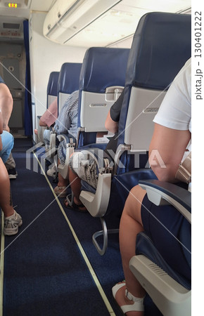 Passengers are seated in an airplane cabin, some relaxing while others check their belongings. The atmosphere is calm as the flight prepares for departure on a sunny afternoon 130401222