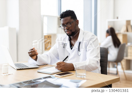 Male African doctor mid-30s sitting at desk writing prescription examining medication, office setting. Professional medical professional working healthcare tasks indoors. 130401381