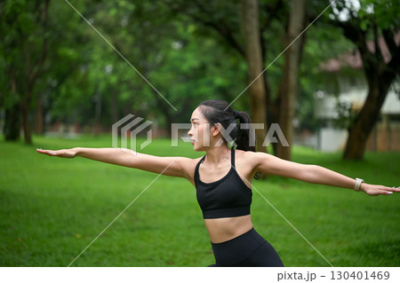 Young woman practicing yoga outdoors, performing warrior pose in a green park 130401469