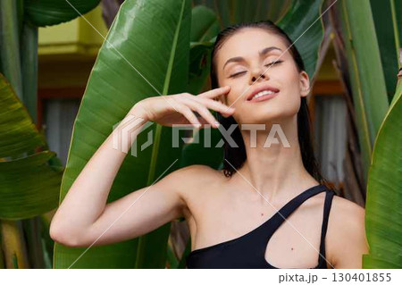 Attractive woman in black swimsuit posing in front of lush green plant with hand on face 130401855