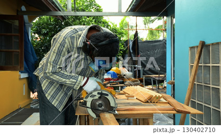 Carpenter using circular saw to cut wooden plank on workbench while wearing safety mask and gloves Carpenter using circular saw to cut wooden plank on workbench while wearing safety mask and gloves 130402710