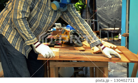 Carpenter aligning wooden planks on workbench while wearing safety mask and gloves with tools in background 130402711