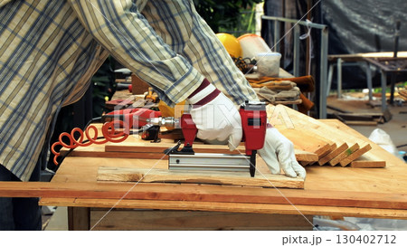 Close up of carpenter hand wearing gloves using red nail gun to fasten wooden plank on workbench in workshop 130402712