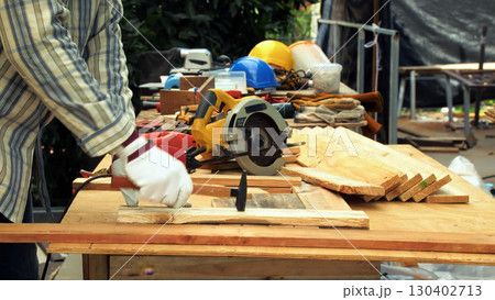 Carpenter hand wearing gloves hammering wooden plank on workbench with tools and safety helmets in background 130402713