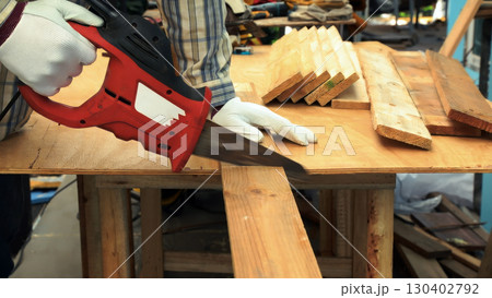 Worker cutting wooden plank with red electric saw on workbench wearing white gloves and striped shirt 130402792