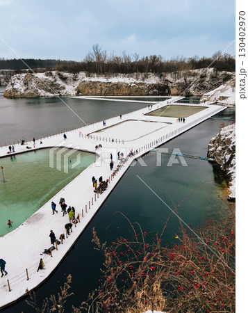 Tourists walking on snowy pier leading to turquoise lagoon surrounded by cliffs covered with snow during winter in Krakow, Poland, creating picturesque winter landscape. Snow covered pier and lagoon 130402970