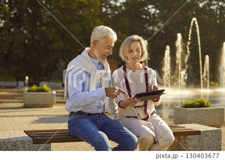 Happy smiling senior couple sitting with digital tablet on the bench in city park. 130403677