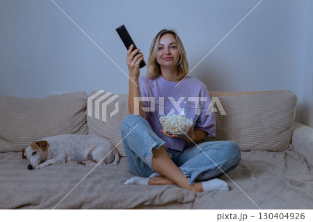 Home, evening: Young woman in casual clothes sits cross-legged on a sofa, holding popcorn and a remote, watching TV with a soft smile. A small dog sleeps peacefully next to her. 130404926