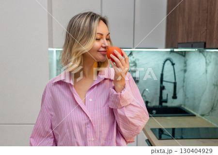 Smiling woman in striped shirt enjoying the scent of a fresh tomato in modern kitchen, eco-friendly vegetables in mesh bag on the table, concept of healthy and organic farm food. Smiling woman in striped shirt enjoying the scent of a fresh tomato in modern kitchen, eco-friendly vegetables in mesh bag on the table, concept of healthy and organic farm food. 130404929