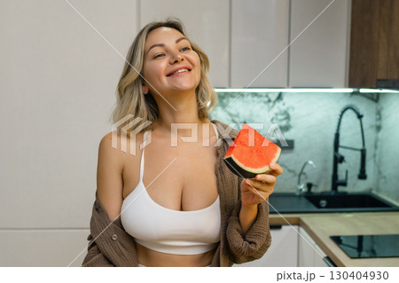 A sensual woman enjoys a slice of juicy watermelon in a modern kitchen. Confident, playful, and radiant, she savors the moment with a hint of summer flirtation. 130404930