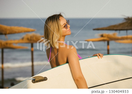 Young blonde woman in a purple bikini holding a surfboard, smiling and enjoying the beach. Ocean waves in the background, summer lifestyle and carefree vacation vibes. 130404933