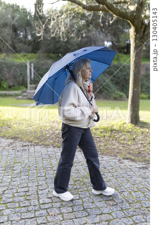 Senior woman walking in park with blue umbrella on cloudy day Senior woman walking in park with blue umbrella on cloudy day 130405143