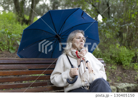 Pensive senior woman holding umbrella sitting on park bench on rainy day 130405150