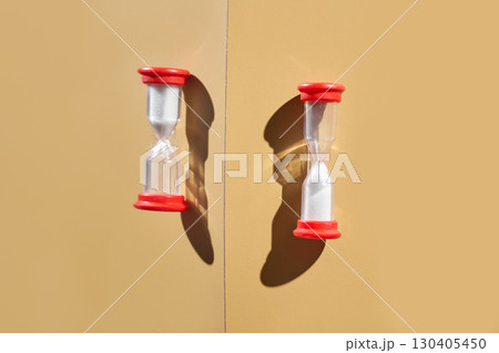 Two red-capped hourglasses casting shadows on a beige background with sunlight 130405450