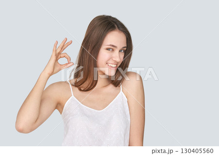 Smiling young caucasian female making ok gesture in white top against light Smiling young caucasian female making ok gesture in white top against light 130405560
