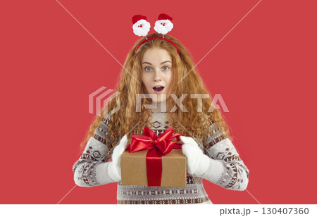 Surprised shocked redhead girl holding Christmas gift present in hands on red background. 130407360