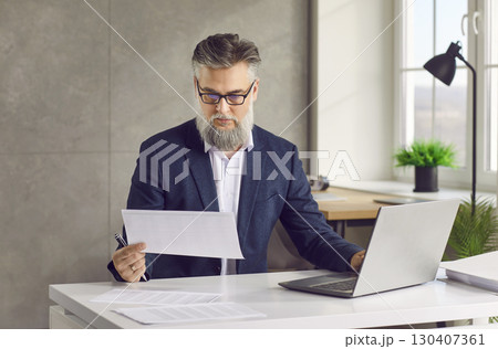 Mature businessman working with paper document and using laptop in his office 130407361