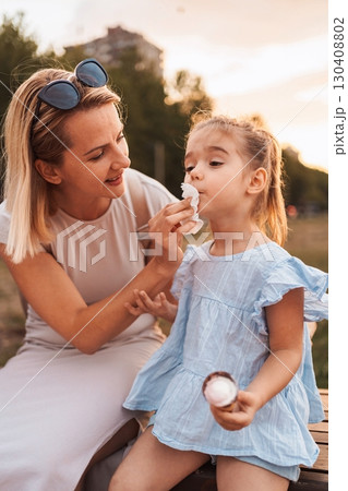 Mother cleaning her daughter's mouth with a napkin after eating ice cream in a park 130408802
