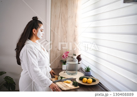 Young woman wearing bathrobe and face mask preparing detox water with lemon and cucumber in the kitchen 130408889