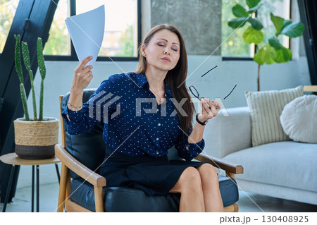 Mature woman experiencing menopausal fever symptoms, waving papers 130408925