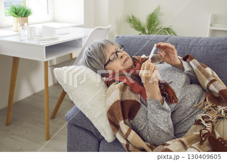 Sick elderly woman who has cold and fever takes her temperature while lying on sofa 130409605
