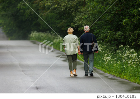Elderly couple holding hands while walking along a quiet country road surrounded by greenery Elderly couple holding hands while walking along a quiet country road surrounded by greenery 130410185