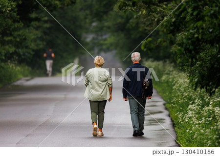 Elderly couple enjoys a leisurely stroll down a quiet, tree-lined pathway in the early morning light Elderly couple enjoys a leisurely stroll down a quiet, tree-lined pathway in the early morning light 130410186