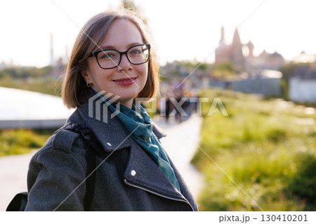 Woman smiles while walking on a pathway surrounded by greenery and city skyline during golden hour 130410201