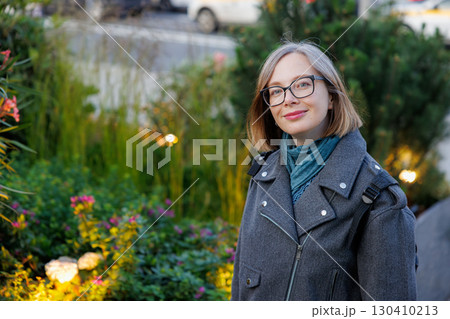Smiling woman poses in a green urban garden on a sunny day while wearing stylish glasses and a warm jacket 130410213