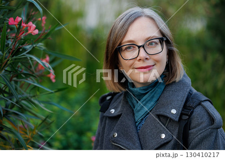 Woman in a gray coat enjoys a blooming garden with pink flowers in a sunny environment during autumn Woman in a gray coat enjoys a blooming garden with pink flowers in a sunny environment during autumn 130410217