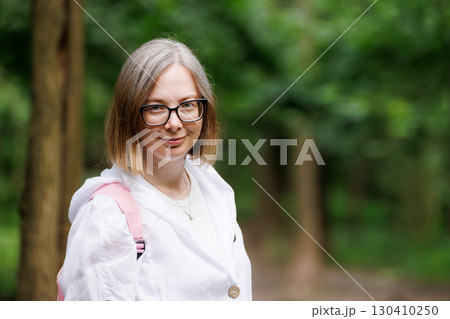 Young woman smiles in a wooded area wearing a white jacket and glasses with a pink backpack on a sunny day Young woman smiles in a wooded area wearing a white jacket and glasses with a pink backpack on a sunny day 130410250