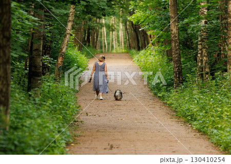 Woman walks pet dog along a peaceful forest path surrounded by lush green trees in summer 130410254