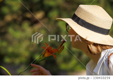 Woman enjoying a warm day in nature while holding a vibrant flower near a blooming garden 130410259