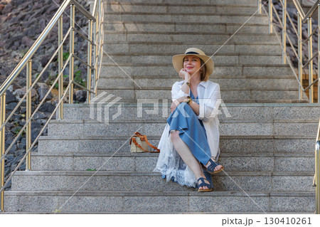 Stylish woman sitting on stone stairs with a wide-brimmed hat during a sunny day in a vibrant urban setting 130410261